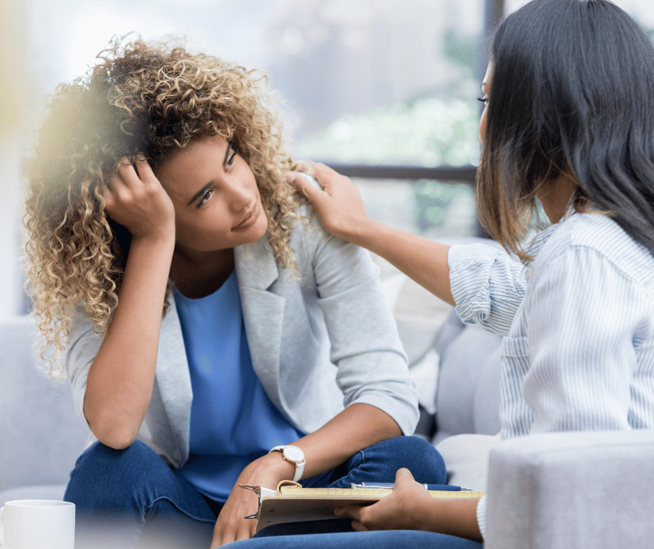 A woman with curly hair looks distressed while sitting on a couch, as another woman, wearing a striped shirt, comforts her by touching her shoulder and speaking to her, with a clipboard in hand.